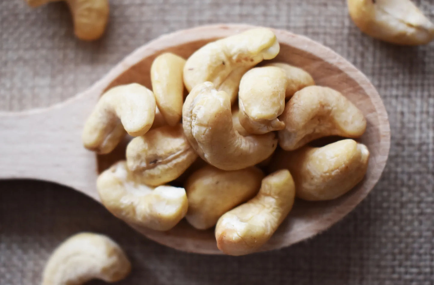 Anacardi naturali interi ammucchiati su un cucchiaio di legno, con altri anacardi sparsi su sfondo in tessuto grezzo — frutta secca premium di qualità Torti Frutta Secca