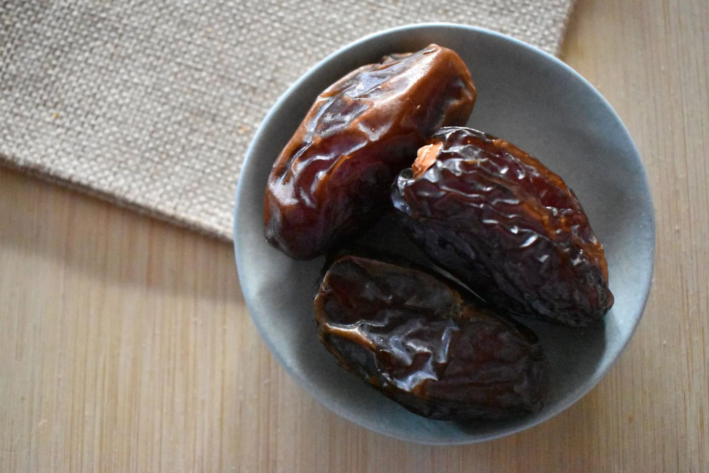 Quattro datteri Medjoul grandi e lucidi in una ciotola di ceramica grigia su superficie in legno chiaro con tovaglietta in juta — frutta secca premium naturale Torti Frutta Secca