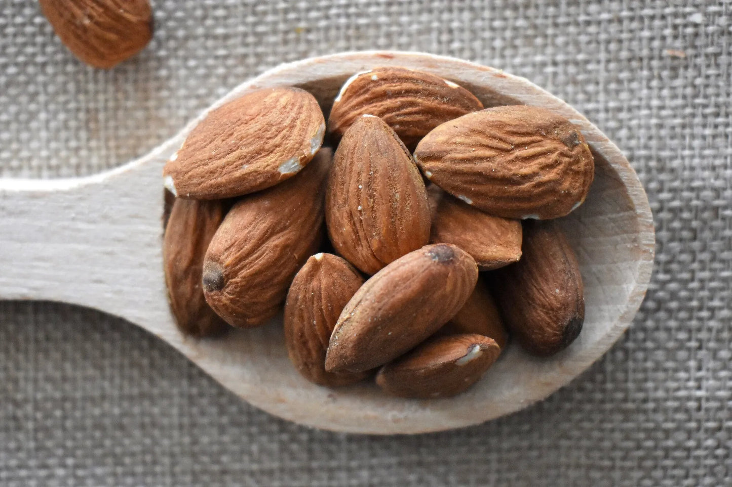 Mandorle naturali sgusciate ammassate su cucchiaio di legno rustico su sfondo di tela di juta grigia — frutta secca premium Torti Frutta Secca