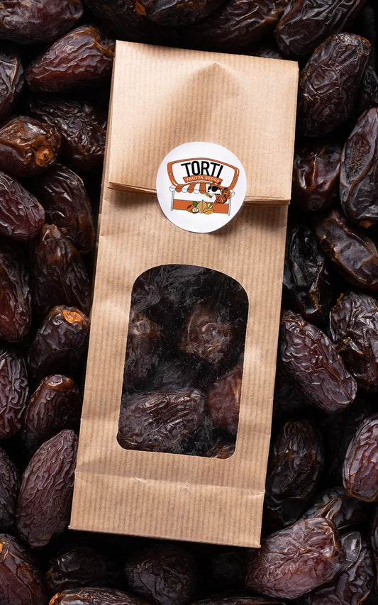 Confezione in carta kraft con finestra trasparente di datteri Medjoul premium Torti Frutta Secca, circondata da datteri sfusi marroni scuri succosi — frutta disidratata premium da comprare online