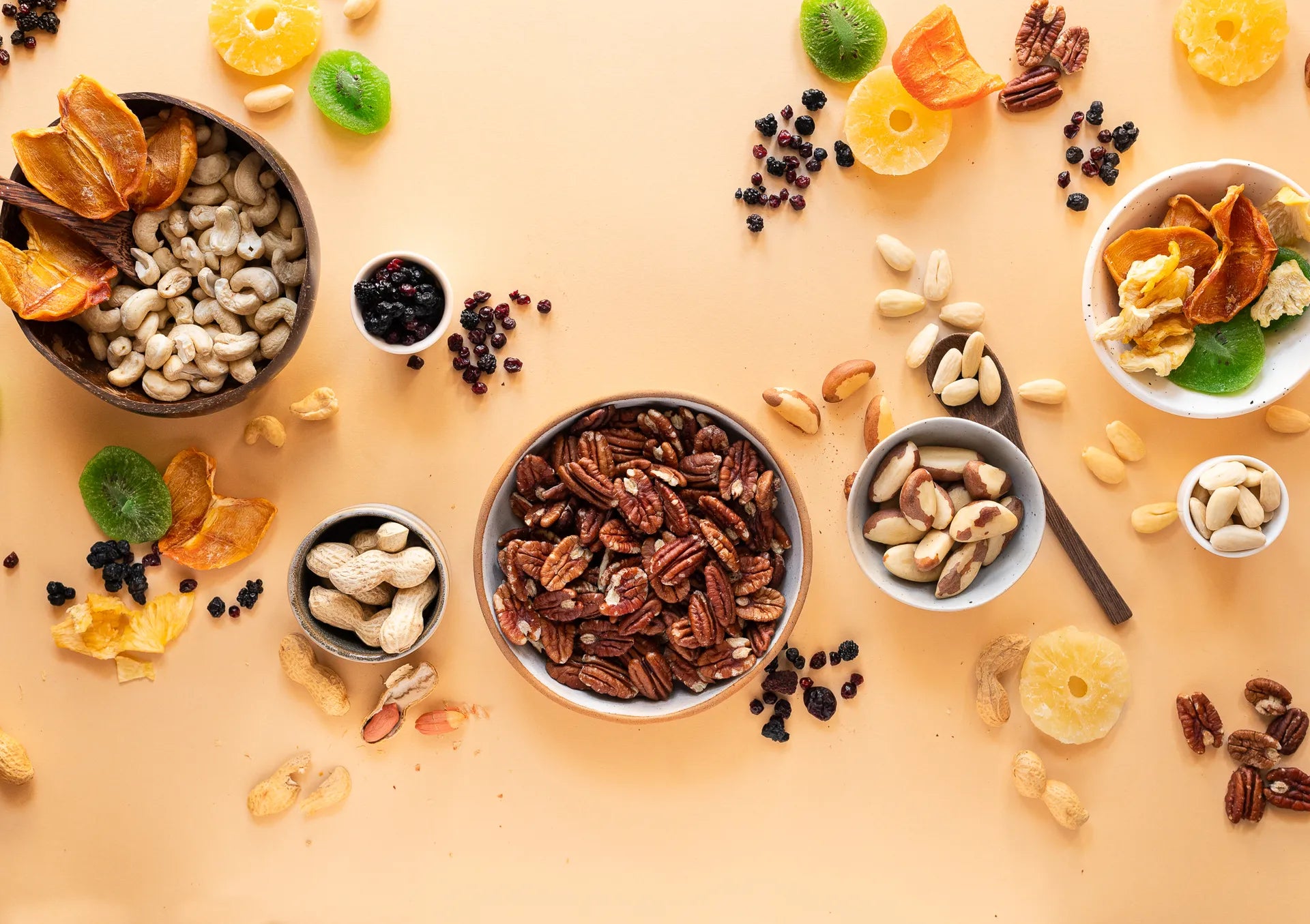 Assortimento di frutta secca premium in ciotole di legno e ceramica: noci pecan, anacardi, arachidi, noci del Brasile, mirtilli essiccati e frutta disidratata come kiwi, albicocche e ananas su sfondo beige — selezione qualità Torti Frutta Secca