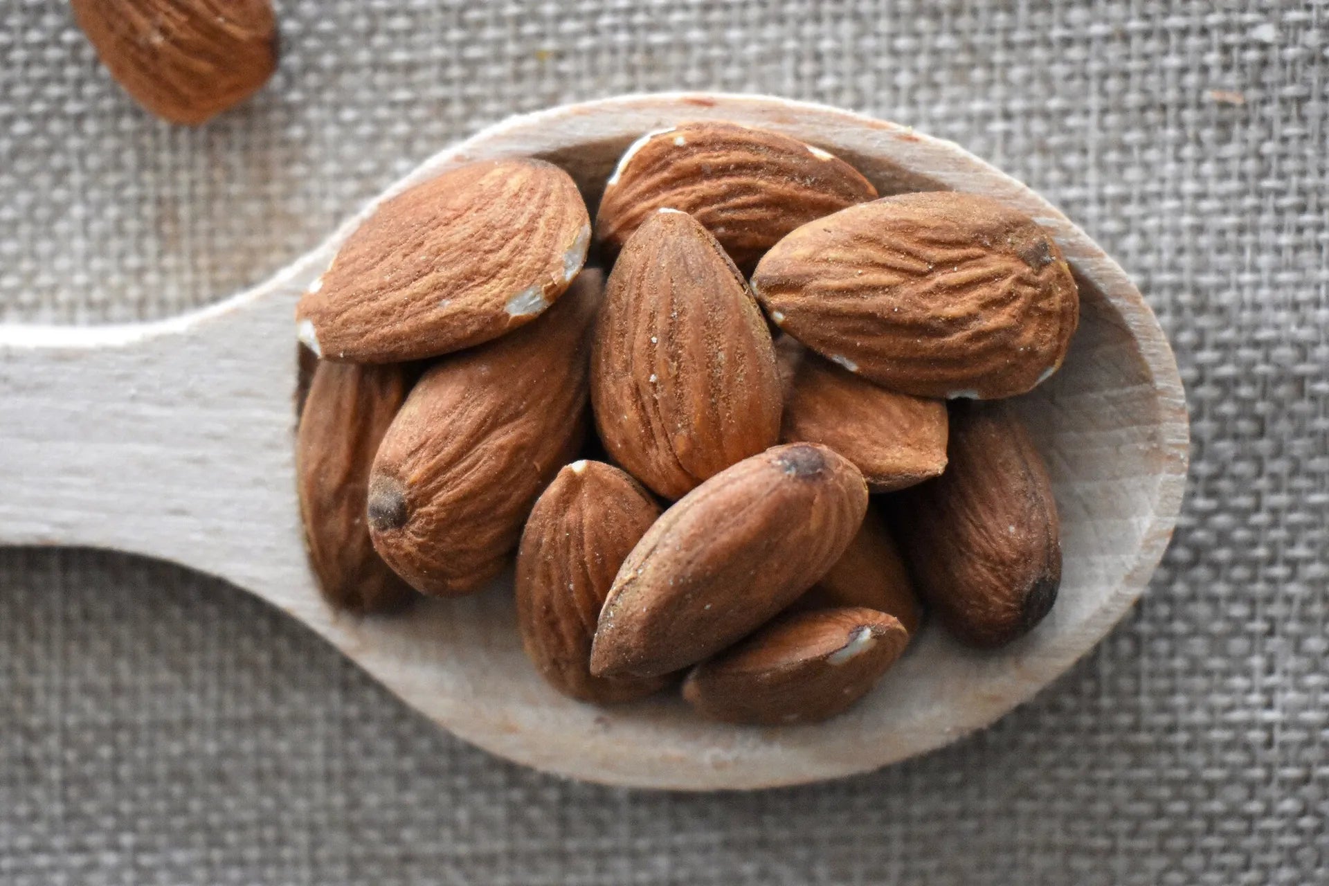 Mandorle naturali sgusciate ammassate su cucchiaio di legno rustico su sfondo di tela di juta grigia — frutta secca premium Torti Frutta Secca