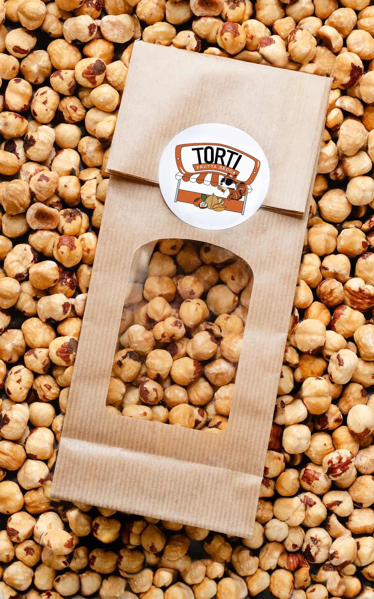 Busta kraft con finestra trasparente piena di nocciole tostate sgusciate Torti Frutta Secca, circondata da nocciole sfuse color avorio e marrone dorato — frutta secca premium acquistabile online