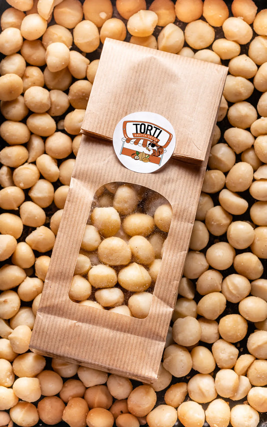 Busta kraft con finestra trasparente di noci macadamia sgusciate Torti Frutta Secca, adagiata su un letto di noci macadamia naturali color avorio — frutta secca premium acquistabile online