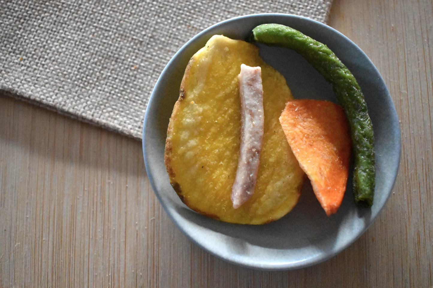 Chips di verdura disidratata premium in ciotola grigia: fetta di patata dorata croccante, striscia di carota arancione e fagiolino verde essiccato, su superficie in legno con tovaglietta in juta