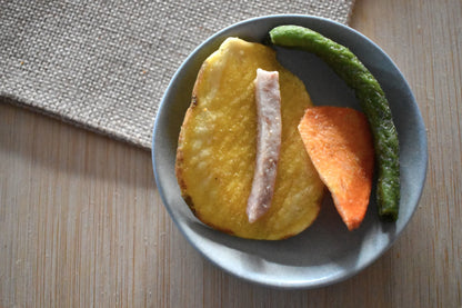 Chips di verdura disidratata premium in ciotola grigia: fetta di patata dorata croccante, striscia di carota arancione e fagiolino verde essiccato, su superficie in legno con tovaglietta in juta